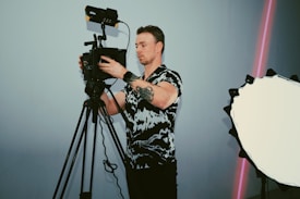 A person is standing behind a professional video camera mounted on a tripod. The individual appears to be adjusting the camera settings. A large reflector with a white surface and black edges is positioned to the right side. The person has a tattoo on their arm and is wearing a patterned shirt. There is a subtle pink light stripe in the background.
