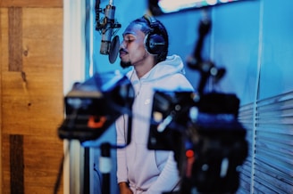 A person wearing headphones and a hoodie is singing or speaking into a professional microphone in a recording studio. The setting includes wooden paneling and blue lighting, giving a modern and focused atmosphere.
