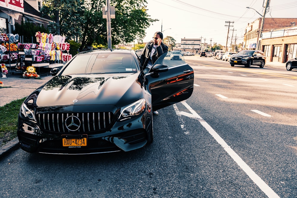 Hombre con chaqueta negra de pie junto a un coche BMW negro en la carretera durante el día