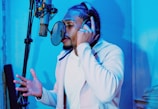 A person with braided hair is wearing headphones and singing or speaking into a microphone. The room is illuminated with blue lighting, creating a modern and cool atmosphere. The individual seems focused and engaged in the activity.