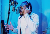 A person with braided hair is wearing headphones and singing or speaking into a microphone. The room is illuminated with blue lighting, creating a modern and cool atmosphere. The individual seems focused and engaged in the activity.