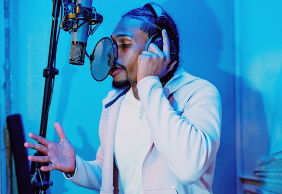 A person with braided hair is wearing headphones and singing or speaking into a microphone. The room is illuminated with blue lighting, creating a modern and cool atmosphere. The individual seems focused and engaged in the activity.