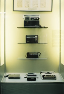 A museum display case contains vintage electronic devices arranged on shelves. The top shelf features a classic radio, followed by another older style radio on the second shelf. The third shelf holds a compact radio, and the bottom shelf displays several gadgets including a typewriter and an early calculator. Light is softly illuminating the display, giving it a nostalgic ambiance.