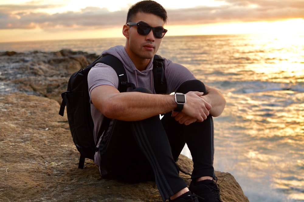 man in black and white polo shirt wearing black sunglasses sitting on brown rock during daytime