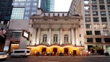 An ornate theater building with neoclassical architecture featuring grand columns and detailed stonework, flanked by modern glass and stone buildings. The entrance is illuminated with warm lights, and a marquee displays show information. People can be seen gathering near the entrance.