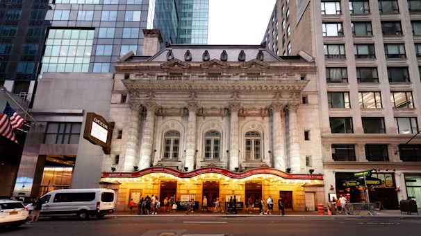 An ornate theater building with neoclassical architecture featuring grand columns and detailed stonework, flanked by modern glass and stone buildings. The entrance is illuminated with warm lights, and a marquee displays show information. People can be seen gathering near the entrance.