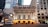 An ornate theater building with neoclassical architecture featuring grand columns and detailed stonework, flanked by modern glass and stone buildings. The entrance is illuminated with warm lights, and a marquee displays show information. People can be seen gathering near the entrance.