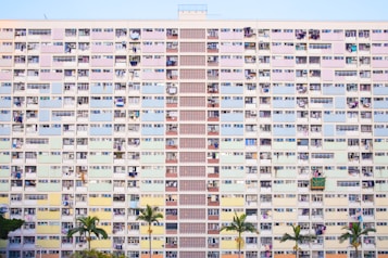A large, multistory apartment building with colorful pastel panels on each unit. The facade features a mix of windows, some open and others closed, along with multiple balconies displaying various personal items like clothes, air conditioning units, and plants. At the base, a few green palm trees stand in front of the building.