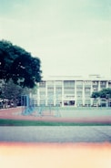 A school playground with a running track and blue exercise structures. In the background is a three-story building with large windows, surrounded by trees. The sky is overcast.