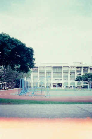 A school playground with a running track and blue exercise structures. In the background is a three-story building with large windows, surrounded by trees. The sky is overcast.