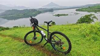 A bicycle with a helmet resting on a lush green hill overlooking a calm lake. The background features a misty landscape with mountains and a serene sky, creating a peaceful atmosphere.