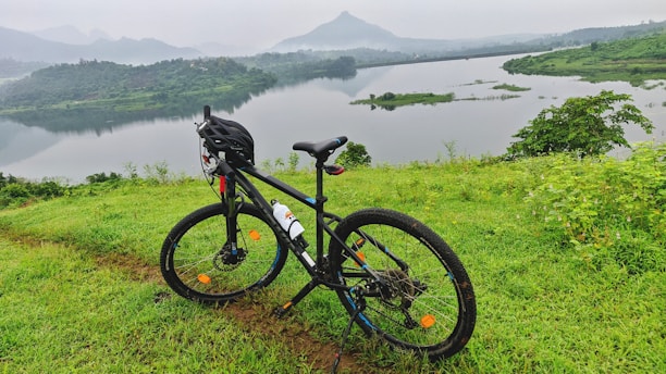A bicycle with a helmet resting on a lush green hill overlooking a calm lake. The background features a misty landscape with mountains and a serene sky, creating a peaceful atmosphere.