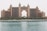 A large, luxurious hotel with a central arch dominates the scene, surrounded by lush greenery and palm trees. The building is an intricate mix of architectural designs with multiple towers and ornate decorations. In the foreground, there is a calm body of water with a rocky shoreline.
