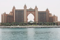 A large, luxurious hotel with a central arch dominates the scene, surrounded by lush greenery and palm trees. The building is an intricate mix of architectural designs with multiple towers and ornate decorations. In the foreground, there is a calm body of water with a rocky shoreline.