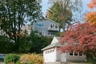 A residential area featuring a light grey two-story house partially obscured by large trees with autumn foliage. The house has a deck with flower pots and a white garage adjacent to it. The area is lush with green, red, and orange leaves, indicating fall season.