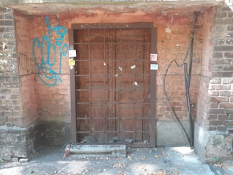 A weathered metal gate is set into a red brick wall, surrounded by peeling plaster and graffiti. The gate appears rusted and is reinforced by a broken piece of wood at the bottom. There is blue graffiti on the left side, and several stickers are attached to the frame.