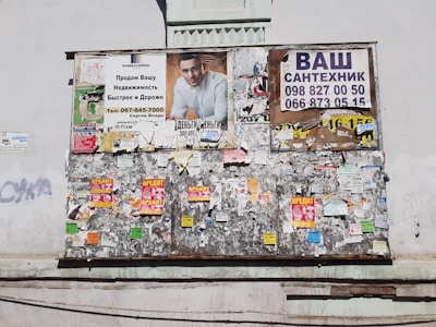 A weathered bulletin board affixed to a wall, covered with a variety of posters and notices. The top section displays larger ads, including an advertisement with a man in a white shirt, and another with contact information for plumbing services. The rest of the board is cluttered with smaller, overlapping papers and flyers in bright colors.