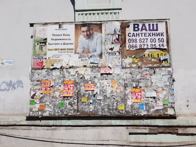 A weathered bulletin board affixed to a wall, covered with a variety of posters and notices. The top section displays larger ads, including an advertisement with a man in a white shirt, and another with contact information for plumbing services. The rest of the board is cluttered with smaller, overlapping papers and flyers in bright colors.