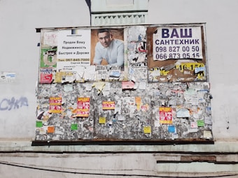 A weathered bulletin board affixed to a wall, covered with a variety of posters and notices. The top section displays larger ads, including an advertisement with a man in a white shirt, and another with contact information for plumbing services. The rest of the board is cluttered with smaller, overlapping papers and flyers in bright colors.