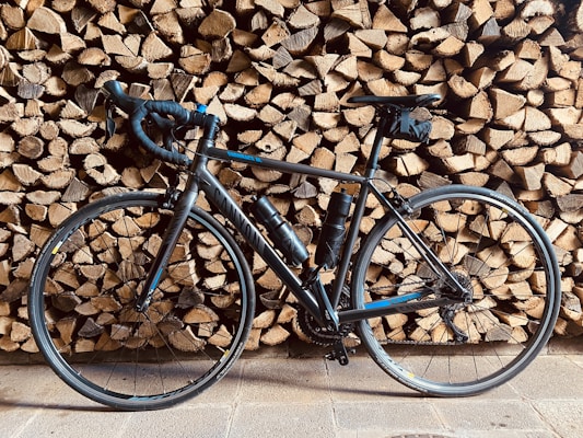 A sleek black road bike is leaning against a stack of neatly arranged firewood. The tires and frame cast shadows on the wooden background, adding depth to the composition. Two water bottles are visible on the bike’s frame, suggesting readiness for long rides.