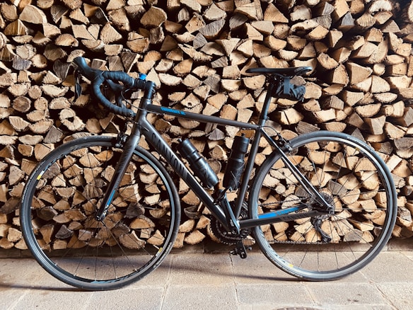 A sleek black road bike is leaning against a stack of neatly arranged firewood. The tires and frame cast shadows on the wooden background, adding depth to the composition. Two water bottles are visible on the bike’s frame, suggesting readiness for long rides.
