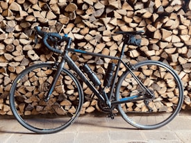 A sleek black road bike is leaning against a stack of neatly arranged firewood. The tires and frame cast shadows on the wooden background, adding depth to the composition. Two water bottles are visible on the bike’s frame, suggesting readiness for long rides.