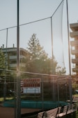 A baseball field with a tall chain-link backstop, a scoreboard, and bright sunlight streaming through trees in an urban setting. Nearby are buildings with several floors, adding to the atmosphere of a city sports area.