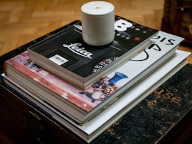 A stack of books is arranged neatly on a dark wooden surface, with a white candle placed on top of the books. The top book has a black cover with the word 'Leica' in white letters and a barcode. The book below features a cover with a Rizzoli publication logo. The surface appears to be a dark wooden table or chest, and the floor is parquet-style wood.