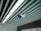 A ceiling-mounted security camera is fixed amidst a series of linear ceiling panels. The panels are aligned in parallel, creating a structured and modern architectural design. The camera is prominent and angled to cover a wide area, indicative of a surveillance setup.