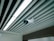A ceiling-mounted security camera is fixed amidst a series of linear ceiling panels. The panels are aligned in parallel, creating a structured and modern architectural design. The camera is prominent and angled to cover a wide area, indicative of a surveillance setup.