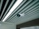 A ceiling-mounted security camera is fixed amidst a series of linear ceiling panels. The panels are aligned in parallel, creating a structured and modern architectural design. The camera is prominent and angled to cover a wide area, indicative of a surveillance setup.