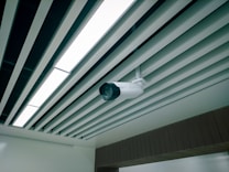 A ceiling-mounted security camera is fixed amidst a series of linear ceiling panels. The panels are aligned in parallel, creating a structured and modern architectural design. The camera is prominent and angled to cover a wide area, indicative of a surveillance setup.