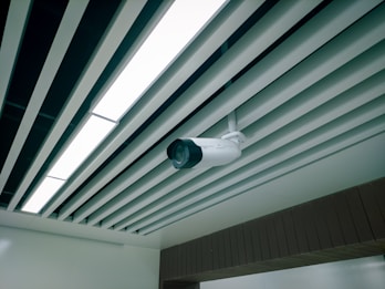 A ceiling-mounted security camera is fixed amidst a series of linear ceiling panels. The panels are aligned in parallel, creating a structured and modern architectural design. The camera is prominent and angled to cover a wide area, indicative of a surveillance setup.