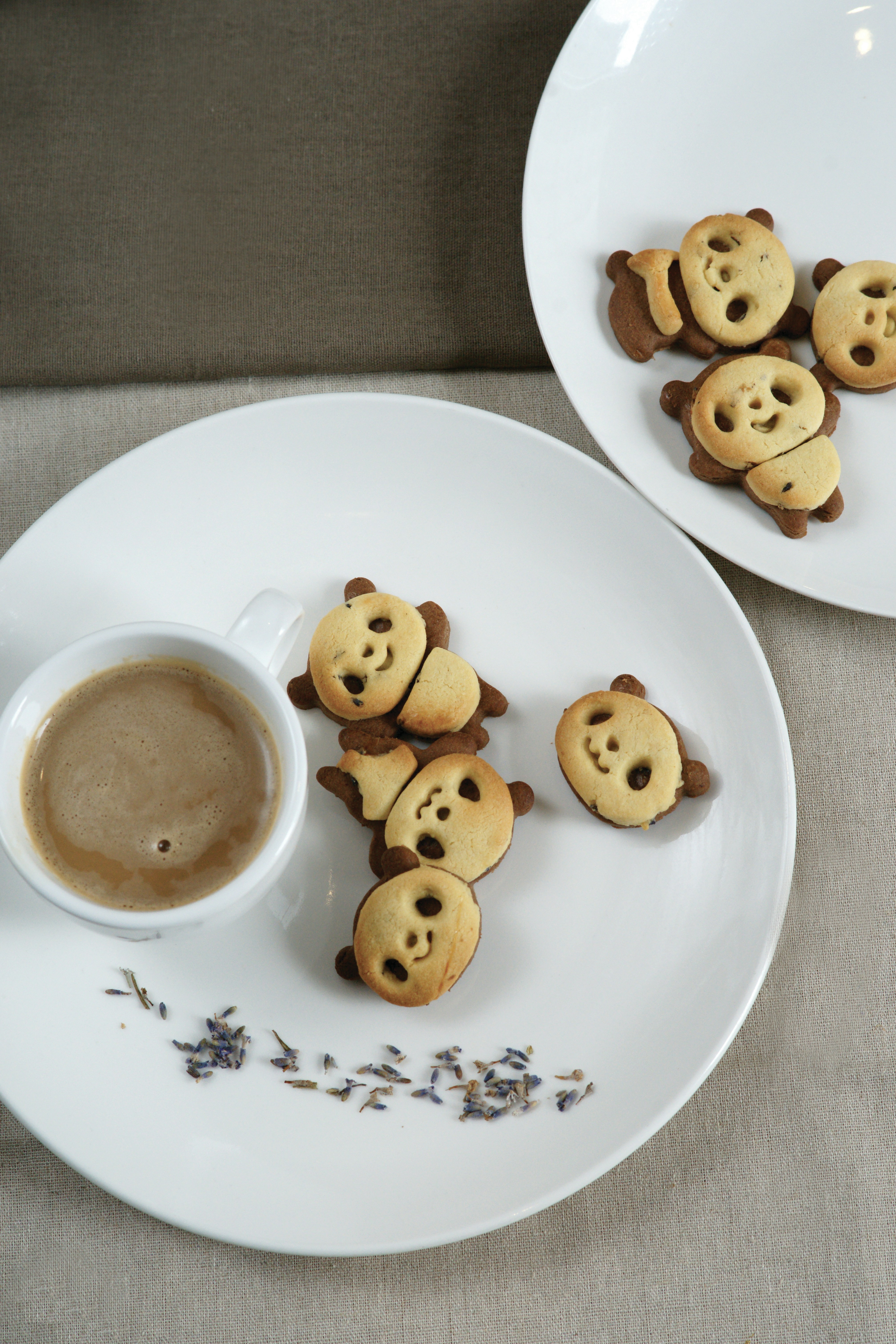 Still-life photograph of panda-shaped cookies surrounding a small cup of coffee on a white plate, with a second plate in the background.