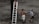Two men are standing near a tall tree on an earthy terrain, with one man holding a ladder while another man with bare torso supports it. A third man is ascending the ladder, with only his legs visible as he climbs. The scene appears casual, with overcast colors and minimal background details.