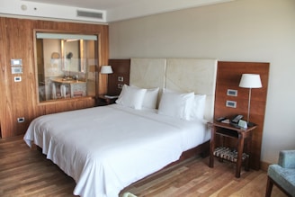 A spacious and modern hotel room with a large, neatly made bed featuring white linens and two plump pillows. The room has wooden floors and a wooden accent wall that includes a large window looking into the bathroom, which has a sink and a mirror. On either side of the bed are bedside tables equipped with lamps, and one has a phone and remote controls. The walls are neutral-colored, creating a calm and relaxing atmosphere.