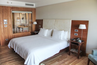 A spacious and modern hotel room with a large, neatly made bed featuring white linens and two plump pillows. The room has wooden floors and a wooden accent wall that includes a large window looking into the bathroom, which has a sink and a mirror. On either side of the bed are bedside tables equipped with lamps, and one has a phone and remote controls. The walls are neutral-colored, creating a calm and relaxing atmosphere.