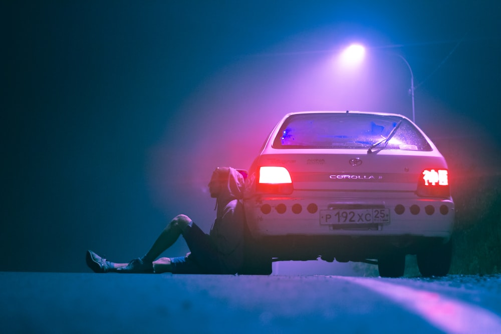 man in black jacket and pants sitting on white car hood during night time