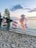 Metallic balloons shaped into the number 25 are displayed on a sandy beach beside a cluster of pink and confetti-filled balloons. The scene is set against a tranquil lakeside with a sunset sky painted with soft clouds.