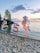 Metallic balloons shaped into the number 25 are displayed on a sandy beach beside a cluster of pink and confetti-filled balloons. The scene is set against a tranquil lakeside with a sunset sky painted with soft clouds.