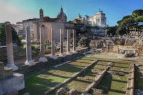 A historic site featuring ancient Roman ruins with tall stone columns in the foreground. In the background, various classical buildings are visible, including a large white structure adorned with statues. The area is surrounded by greenery, and the scene is lit by daylight, casting shadows across the site.