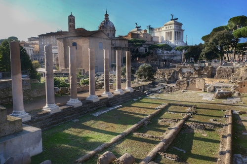 A historic site featuring ancient Roman ruins with tall stone columns in the foreground. In the background, various classical buildings are visible, including a large white structure adorned with statues. The area is surrounded by greenery, and the scene is lit by daylight, casting shadows across the site.