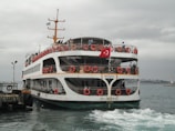 A ferry with red lifebuoys and a Turkish flag is approaching a dock. The sky is overcast, and the water appears slightly choppy. Several people are visible on the ferry, standing on the decks.