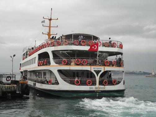 A ferry with red lifebuoys and a Turkish flag is approaching a dock. The sky is overcast, and the water appears slightly choppy. Several people are visible on the ferry, standing on the decks.