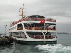 A ferry with red lifebuoys and a Turkish flag is approaching a dock. The sky is overcast, and the water appears slightly choppy. Several people are visible on the ferry, standing on the decks.