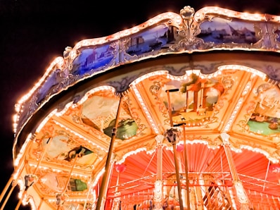 A brightly lit carousel with intricate decorative panels depicting various scenes. The carousel is illuminated by warm lights, creating a lively glow against the dark background.
