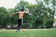 A shirtless person wearing athletic pants and bright blue cleats is about to kick a soccer ball on a grassy field. Surrounding the field are tall, green trees, and there is a soccer goal in the background. The scene conveys movement and energy, with the athlete's arm raised and body positioned in a kicking stance.