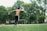 A shirtless person wearing athletic pants and bright blue cleats is about to kick a soccer ball on a grassy field. Surrounding the field are tall, green trees, and there is a soccer goal in the background. The scene conveys movement and energy, with the athlete's arm raised and body positioned in a kicking stance.