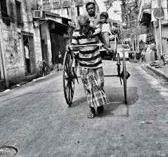 A man pulls a traditional hand-pulled rickshaw through a narrow street, with a passenger and a child sitting in the vehicle. The street is lined with old buildings, and a person stands in a doorway watching the scene. Bicycles and signs indicate a typical urban environment.