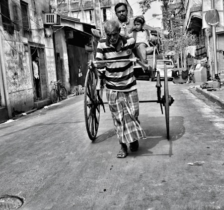 A man pulls a traditional hand-pulled rickshaw through a narrow street, with a passenger and a child sitting in the vehicle. The street is lined with old buildings, and a person stands in a doorway watching the scene. Bicycles and signs indicate a typical urban environment.